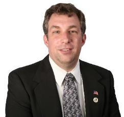 headshot of a person in black blazer, white button up shirt, and patterned tie in front of a white backdrop